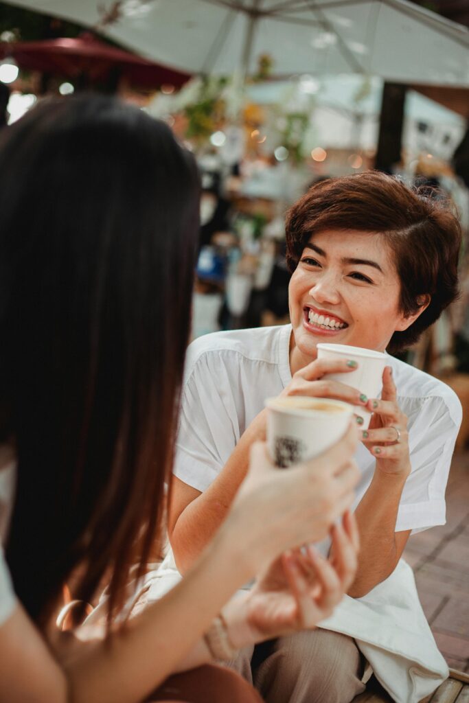 Laughing attractive Asian females enjoying takeaway coffee in modern outdoor coffee shop and looking at each other happily