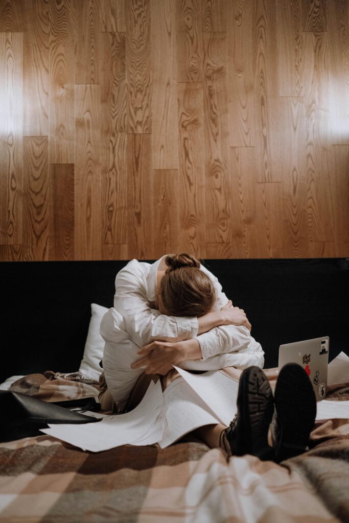 Woman sitting on bed surrounded by papers, indicating stress and exhaustion.